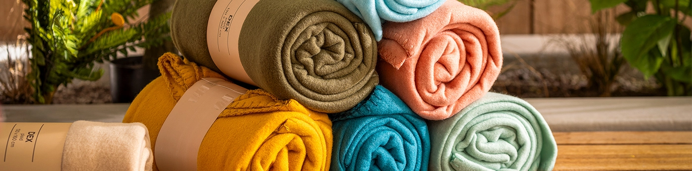 Maak-elke-avond-perfect-met-dutch-decor-terrasplaids Een stapel kleurrijke, opgerolde terrasplaids in groene, blauwe, gele, oranje en beige tinten op een houten bankje voegt een vleugje Dutch Decor toe aan ieder interieur